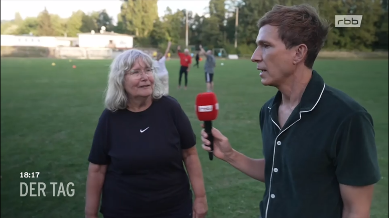 rbb-Reporter Arndt Breitfeld rechts im Bild im Gespräch mit Elke, im Hintergrund auf Rasensportplatz sieht man Menschen beim Sport